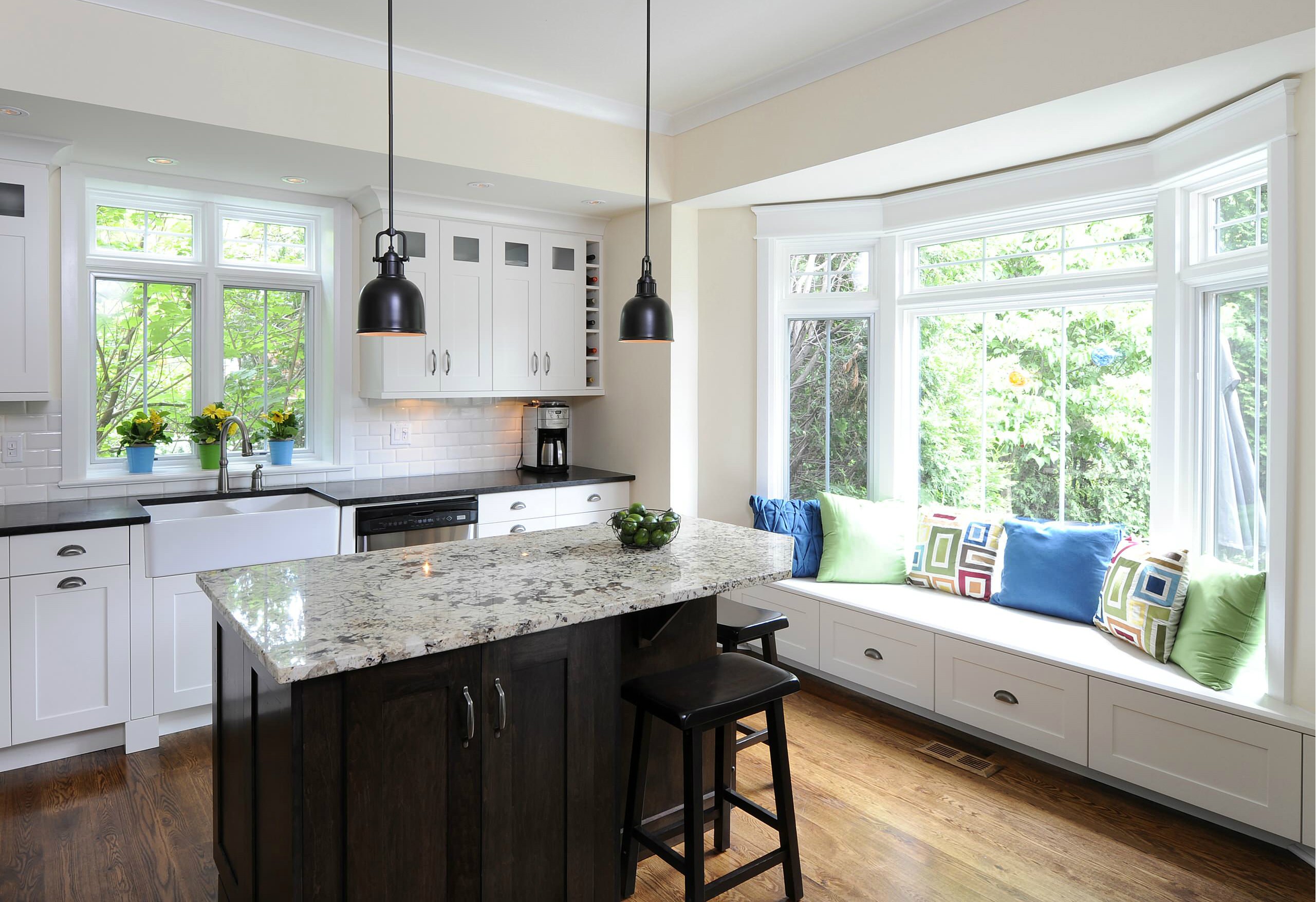 Gorgeous bay window seat with white shaker cabinets and transom windows plus black kitchen island and granite countertops also pendant lighting and wood floor